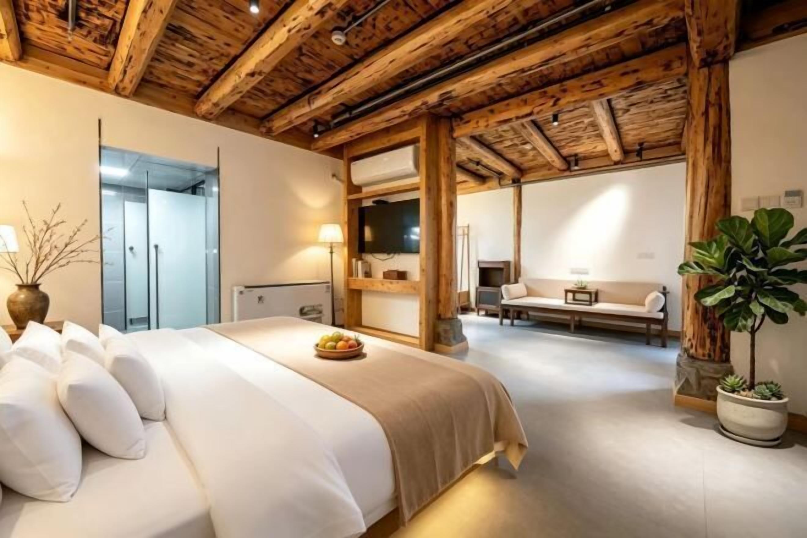 A bedroom with a wooden ceiling, a bed with white bedding, a bedside table, a potted plant, and a view of a bathroom and living area.