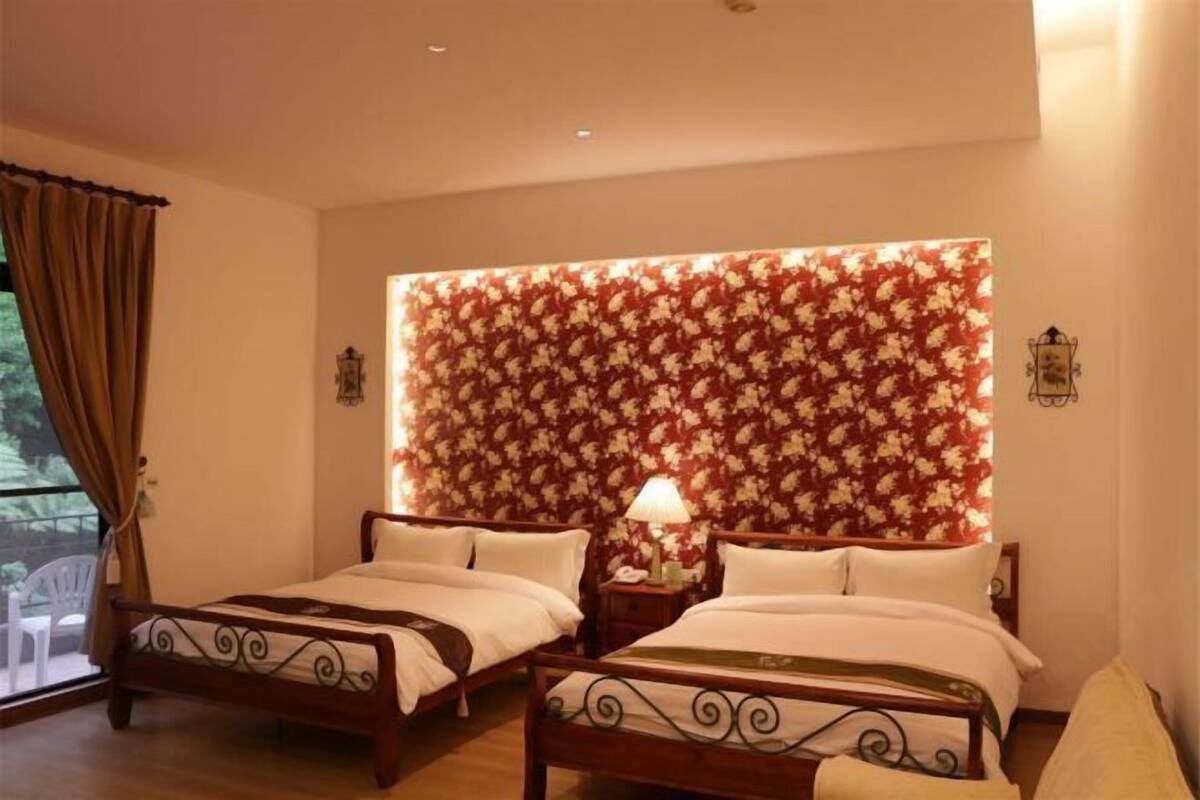 Een hotelkamer met twee bedden, een hoofdbord met bloemenprint, een nachtkastje met lamp en een raam met gordijnen.