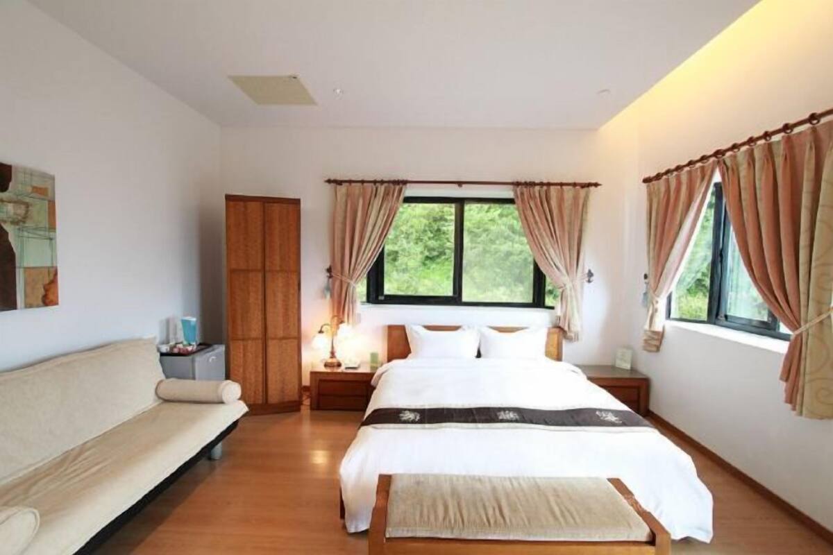 Een slaapkamer met een bed, een nachtkastje, een houten kledingkast, een bank en een raam met gordijnen.