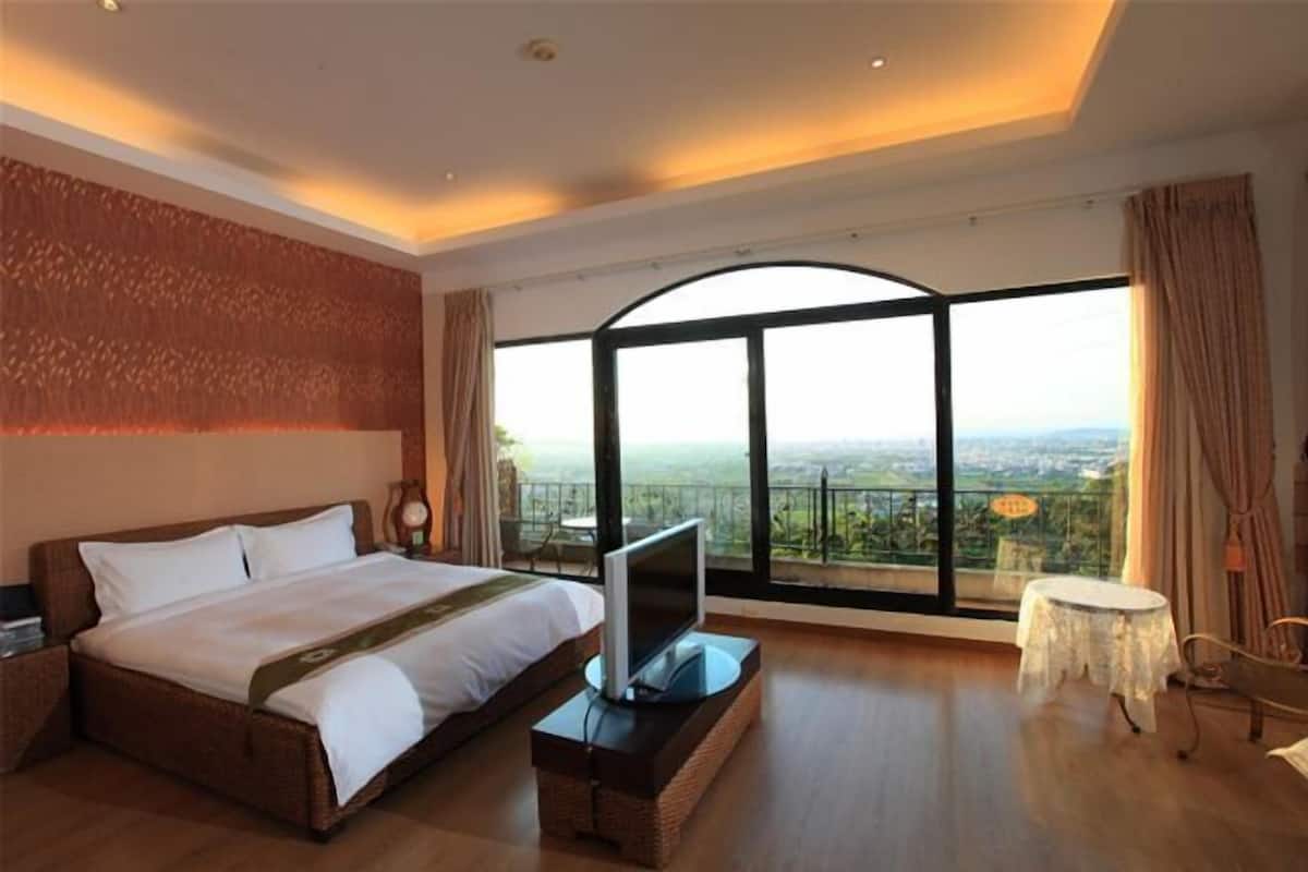 Een hotelkamer met een groot bed, een televisie op een standaard, een klein tafeltje en uitzicht op de stad door grote ramen.