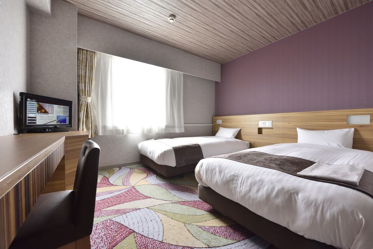 A hotel room with two beds, a desk, a television, and a large window with curtains.