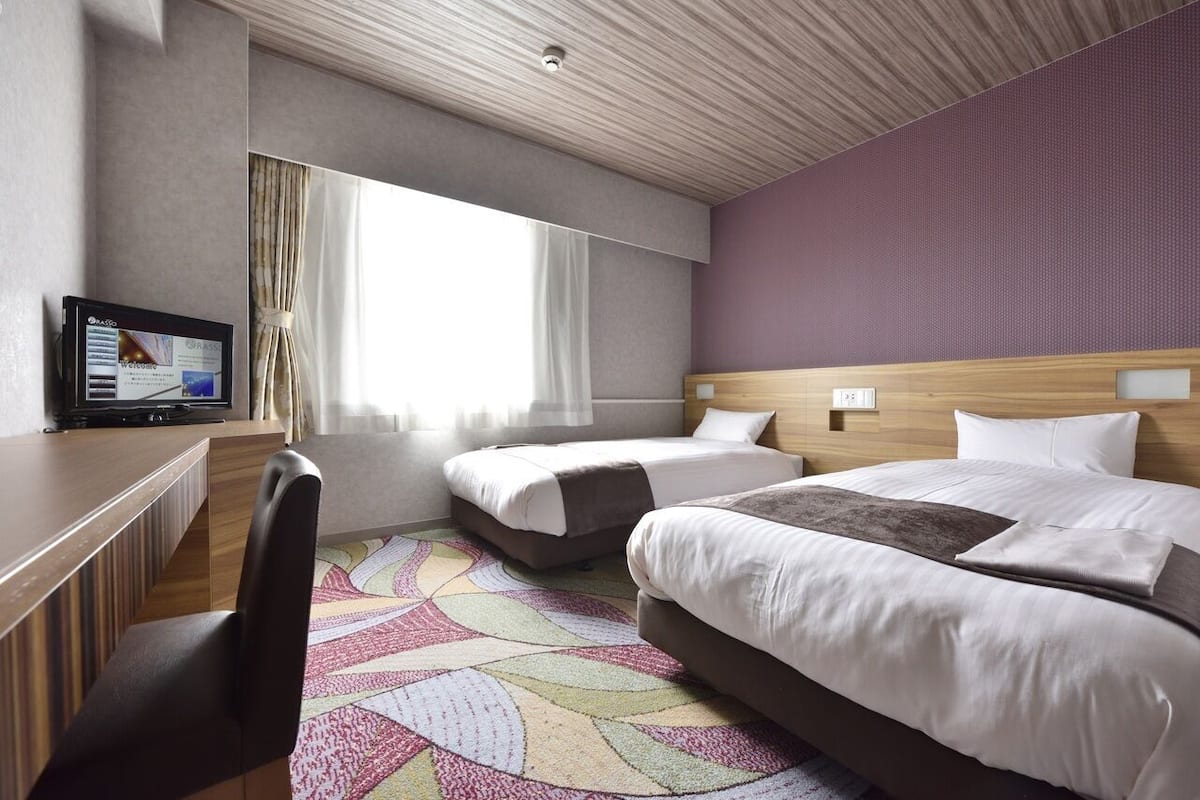 Hotellihuone, jossa on kaksi sänkyä, työpöytä, televisio ja suuri ikkuna, jossa on verhot.