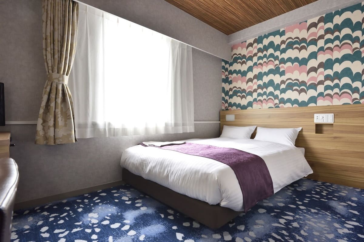 Hotellihuone, jossa on sänky, puinen pääty, ikkuna, jossa on verhot, kuvioitu seinä ja sininen matto.