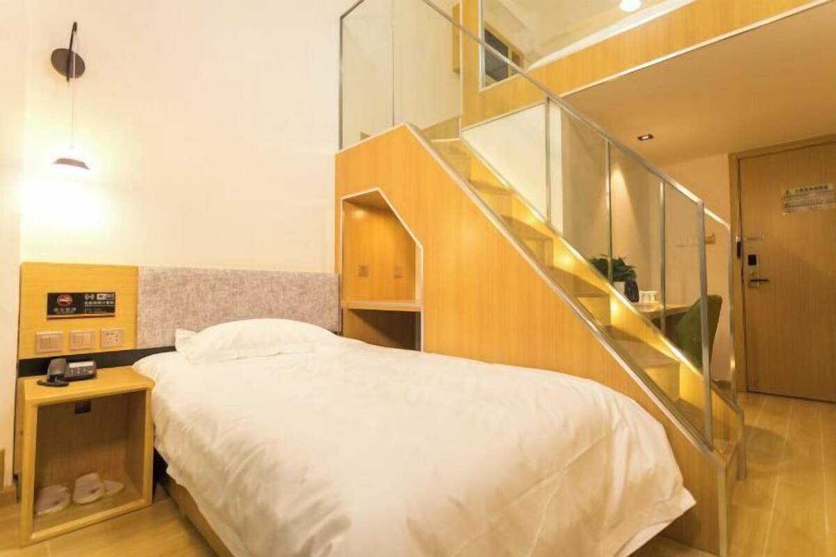 A hotel room with a bed, a nightstand with a phone and a lamp, a wooden staircase, and a door with a sign.