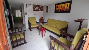 A living room with a yellow sofa, wooden chairs, a red stool, and a painting on the wall.