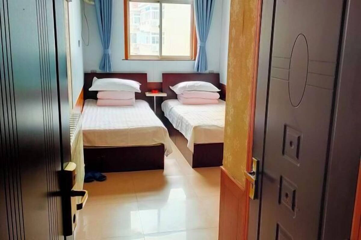 Quarto de hotel com duas camas, uma mesinha, uma janela com cortinas e uma porta.