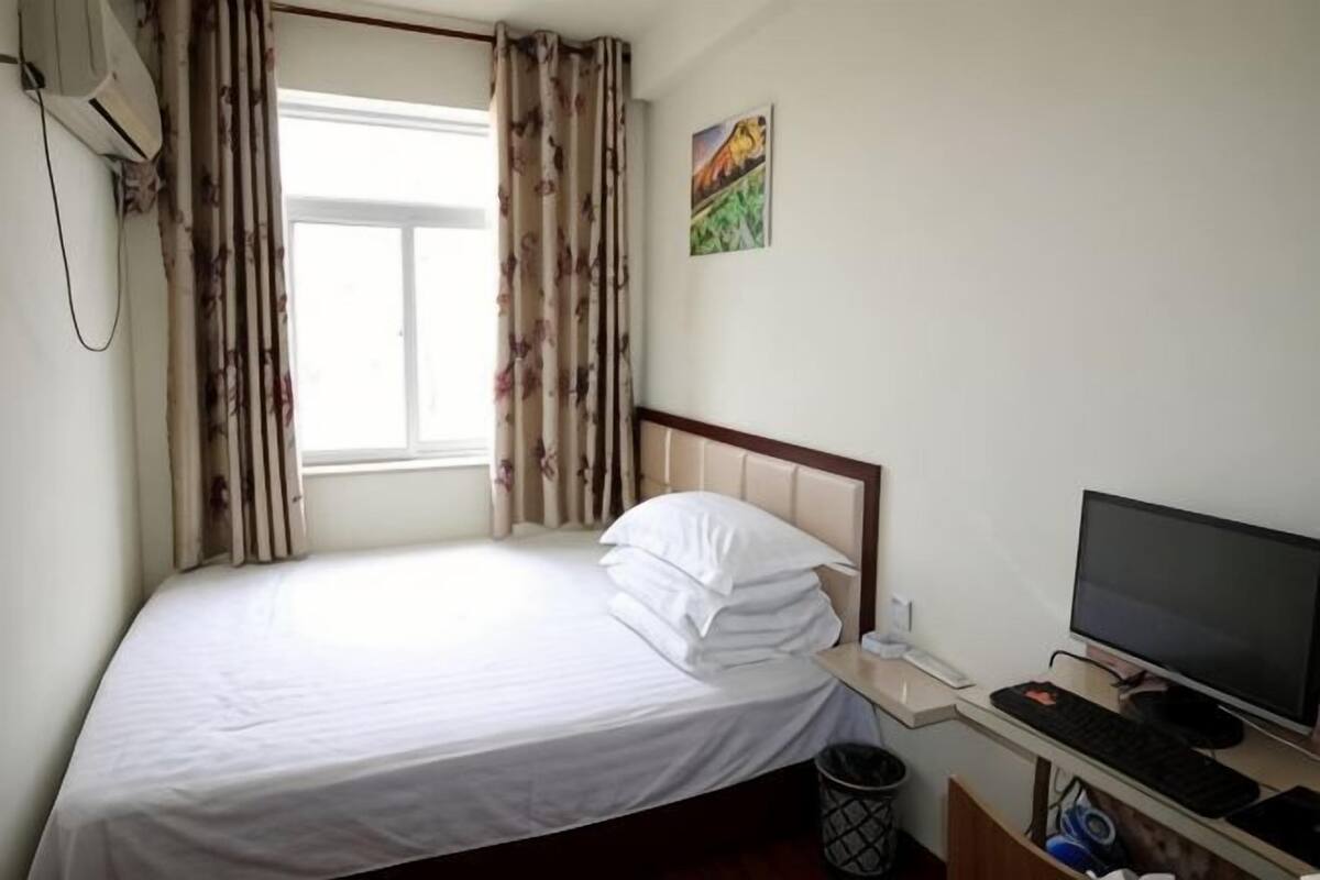 Uma cama de solteiro com roupa de cama branca, uma escrivaninha com um computador e uma janela com cortinas.