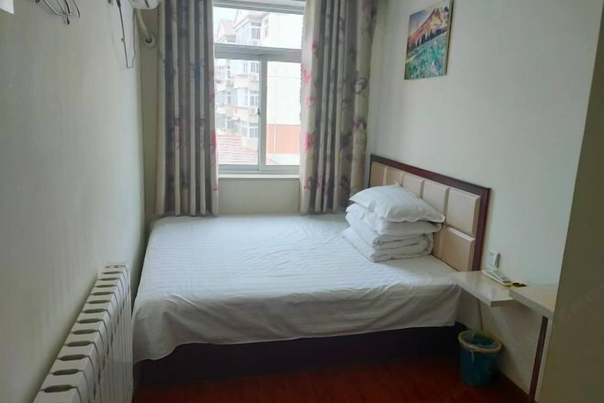 Uma cama de solteiro com roupa de cama branca, um radiador, uma janela com cortinas e uma pequena lixeira.
