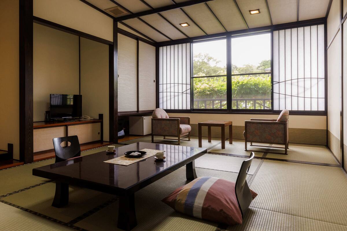 Une pièce de style japonais traditionnel, avec un sol recouvert de tatamis, une table basse, des chaises, un téléviseur et une vue sur la verdure environnante.
