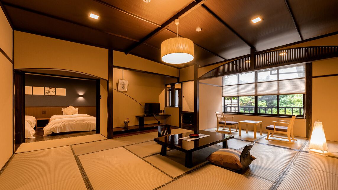 Une pièce de style japonais traditionnel, avec un sol recouvert de tatamis, une table basse avec des coussins, un lit avec une literie blanche et une grande fenêtre donnant sur un paysage verdoyant.