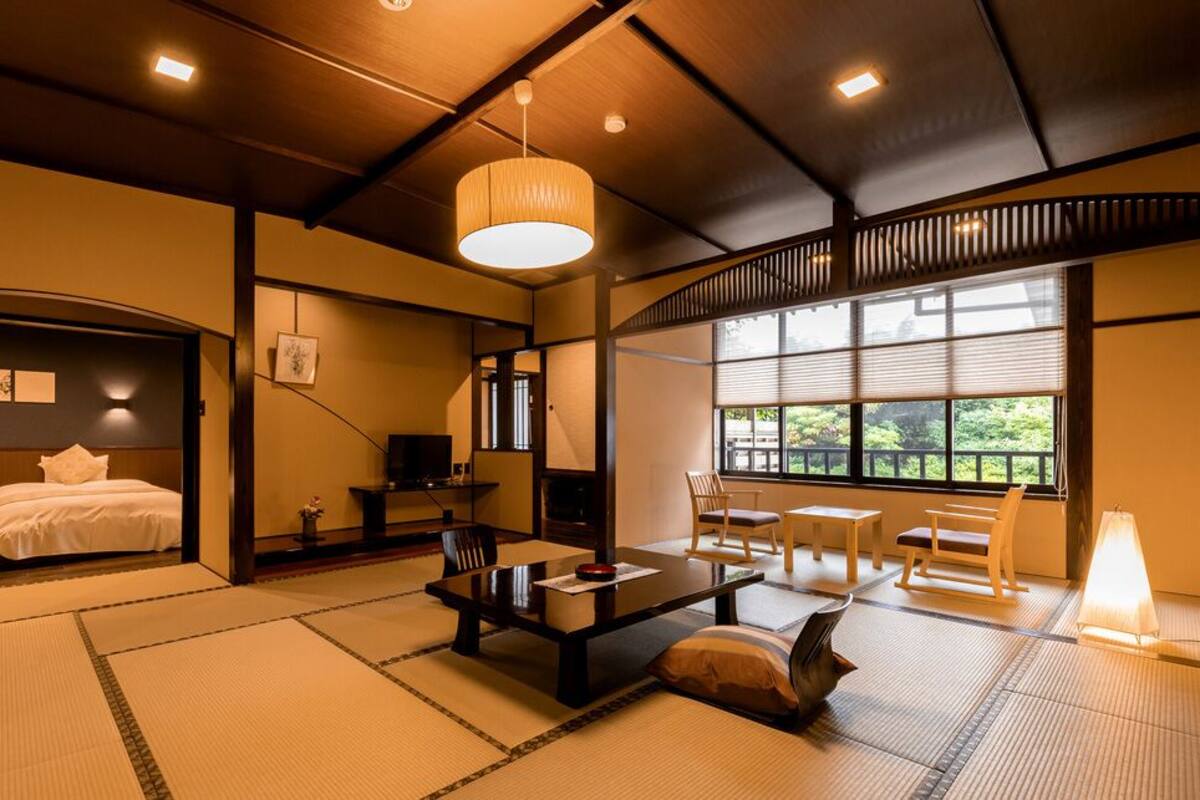 Une pièce de style japonais traditionnel, avec un sol recouvert de tatamis, une table basse avec des coussins, un lit avec une literie blanche et une grande fenêtre donnant sur un paysage verdoyant.