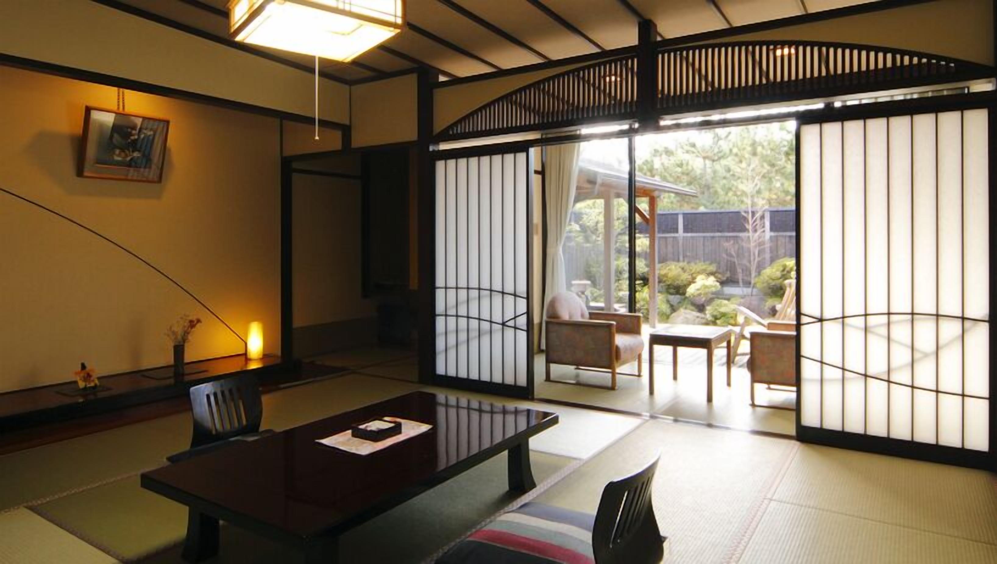 A traditional Japanese room with tatami flooring, a low table, and sliding doors leading to an outdoor area with a view of greenery.