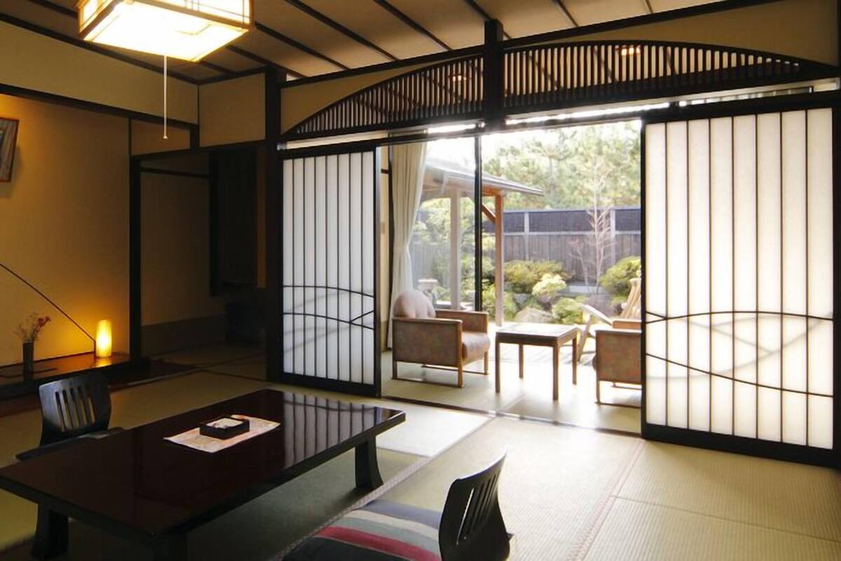Une pièce de style japonais traditionnel, avec un sol recouvert de tatamis, une table basse et des portes coulissantes donnant sur un espace extérieur offrant une vue sur la verdure.