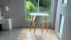 A room with a wooden floor, a white desk, a white chair, and a window with a view of greenery. - Sealoft,Pattaya (Pattaya)