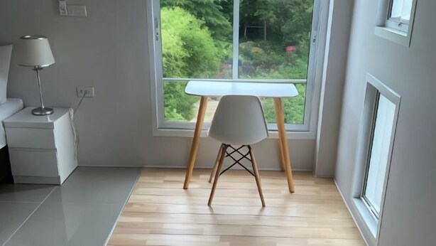 A room with a wooden floor, a white desk, a white chair, and a window with a view of greenery. - Sealoft,Pattaya (Pattaya)