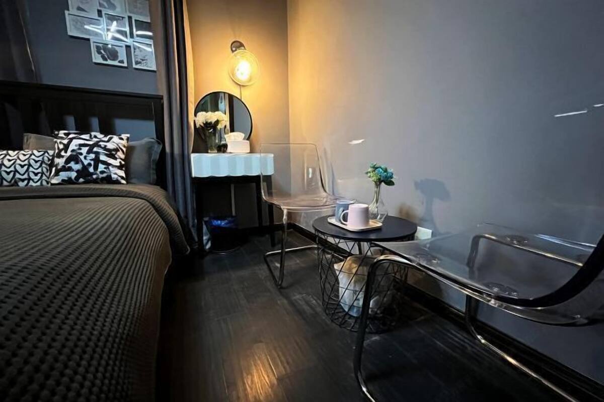 A modern hotel room with a bed, a small table with a cup and a vase, a mirror, and a wall-mounted light.