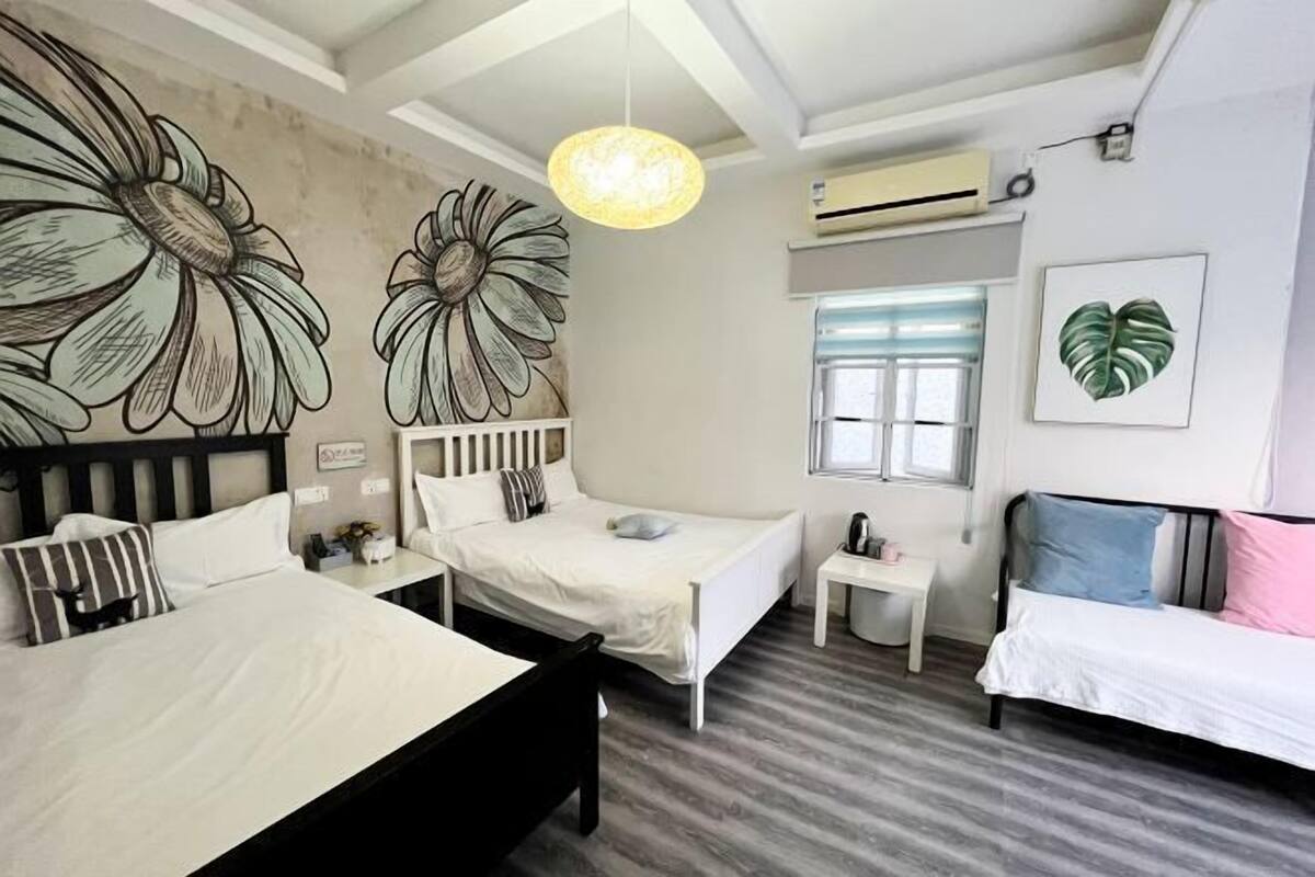 A bedroom with a bed, a bedside table, a chair, a window, and a wall with floral artwork.