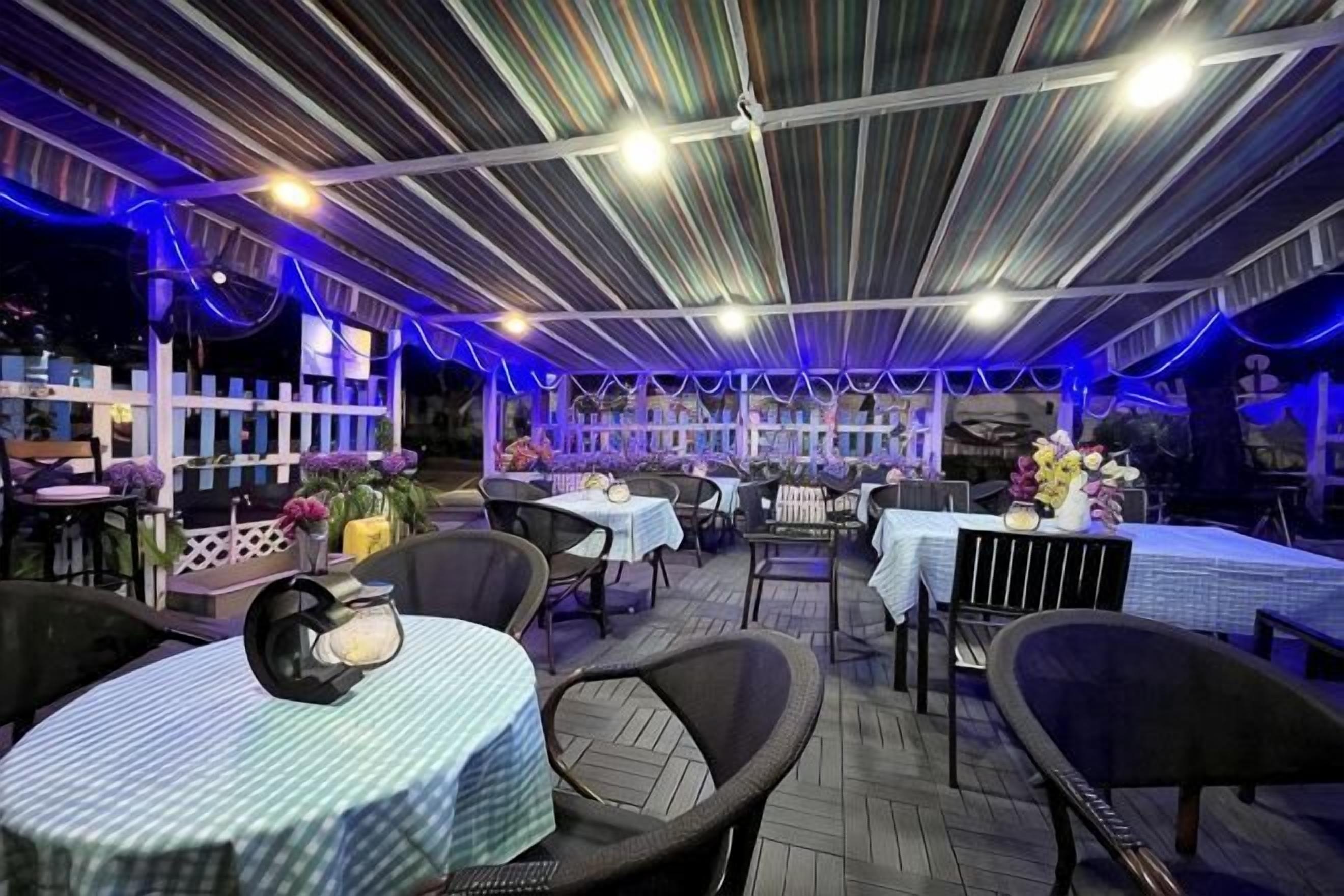 An outdoor dining area with tables and chairs, a covered section with a striped ceiling, and a white fence in the background.