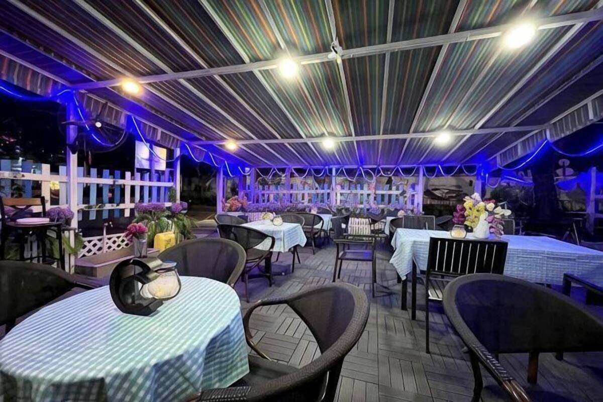 An outdoor dining area with tables and chairs, a covered section with a striped ceiling, and a white fence in the background.