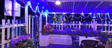 Un patio al aire libre con mesas y sillas, rodeado por una cerca blanca y decorado con luces colgantes.