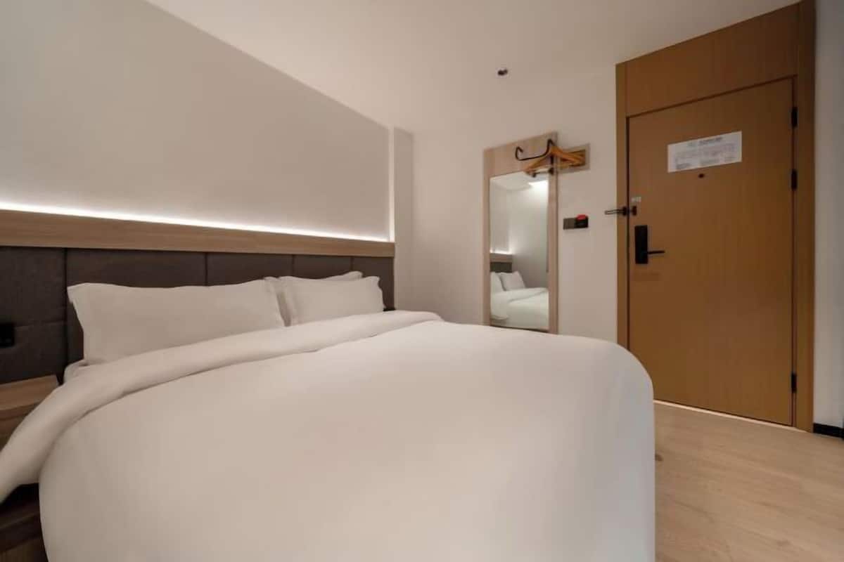 A modern hotel room with a large bed, a wooden headboard, a mirror, and a wooden door.