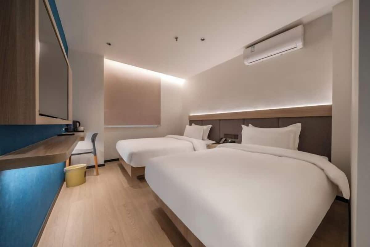A modern hotel room with two single beds, a small desk, a chair, and a window with blinds.
