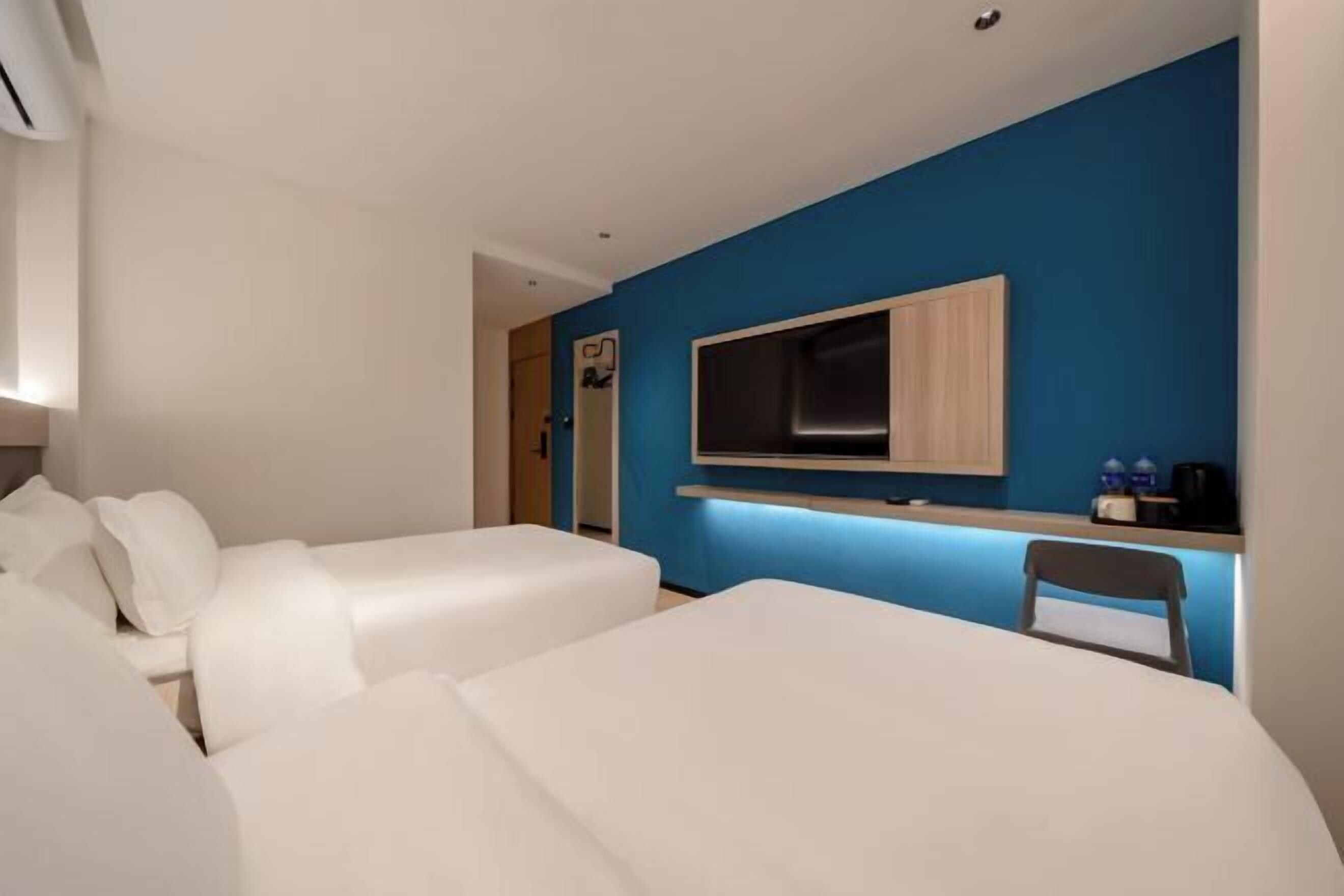 Quarto de hotel moderno com uma parede de destaque azul, uma TV de tela plana fixada acima de um console de madeira, e duas camas brancas.