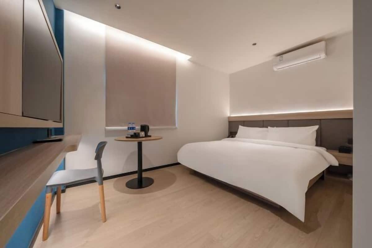 A modern hotel room with a bed, a small round table with a chair, a TV mounted on the wall, and a window providing natural light.