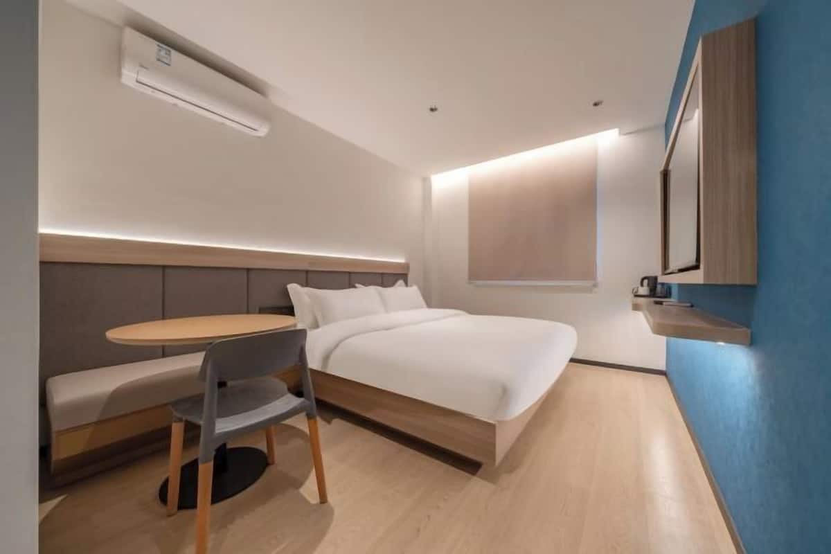 A modern hotel room with a bed, a small table with a chair, a built-in bench, a wall-mounted TV, and an air conditioning unit.