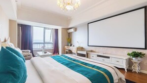 Ein Hotelzimmer mit einem großen Bett, einem Schreibtisch, einem Sessel, einem kleinen Tisch, einer Topfpflanze und einem großen Fenster mit Vorhängen.