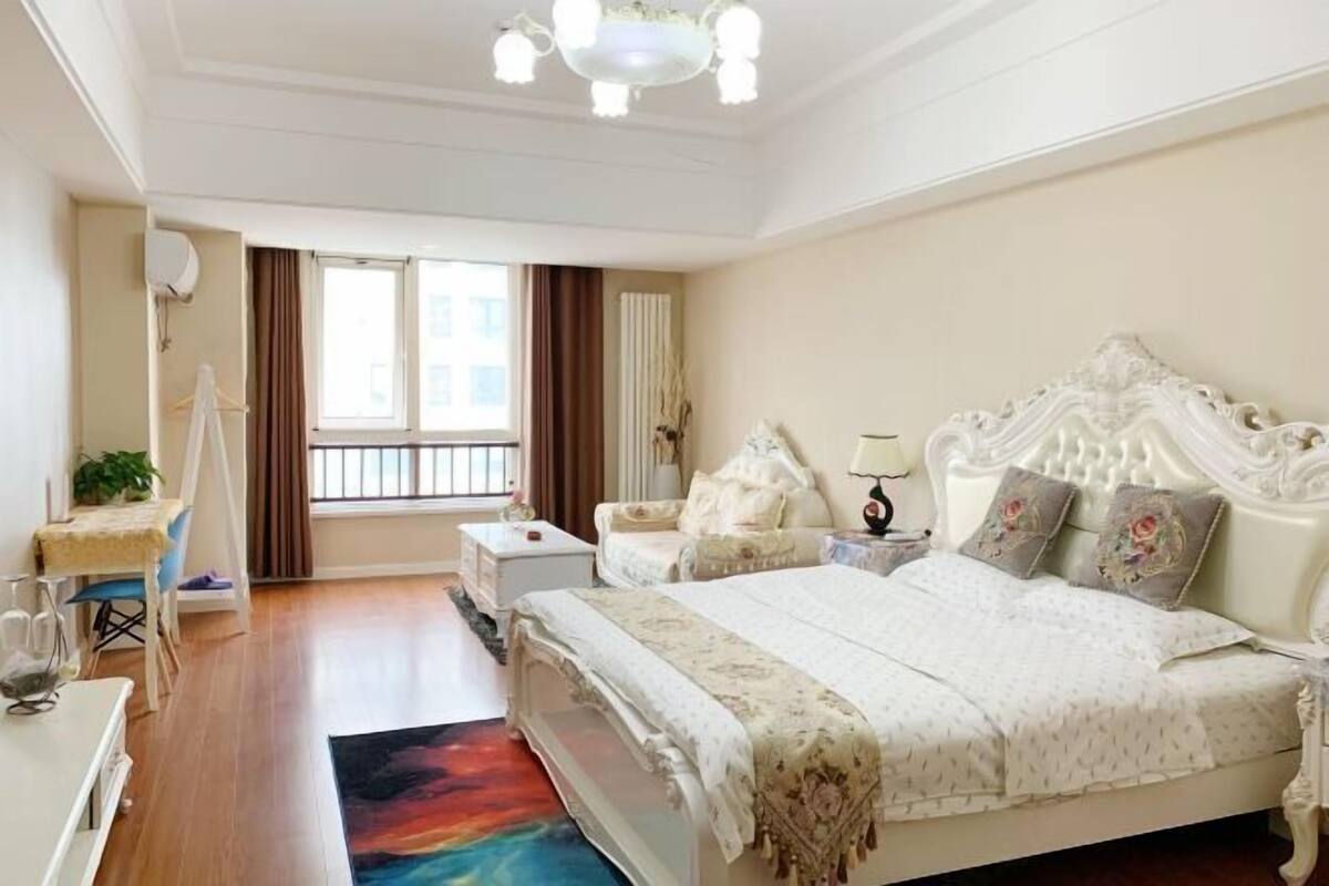 Une chambre à coucher avec un grand lit, une table de chevet, une commode, une coiffeuse, une fenêtre avec des rideaux et un lustre.