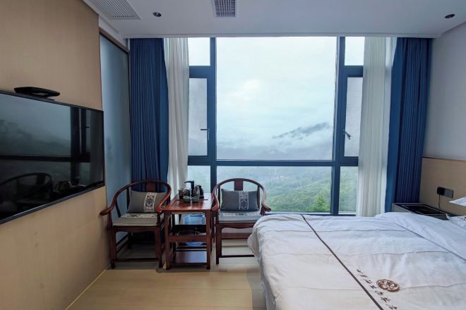 Habitación de hotel con cama, televisión, escritorio con sillas y un gran ventanal con vistas a la montaña.