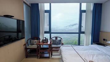 Habitación de hotel con cama, televisión, escritorio con sillas y un gran ventanal con vistas a la montaña.