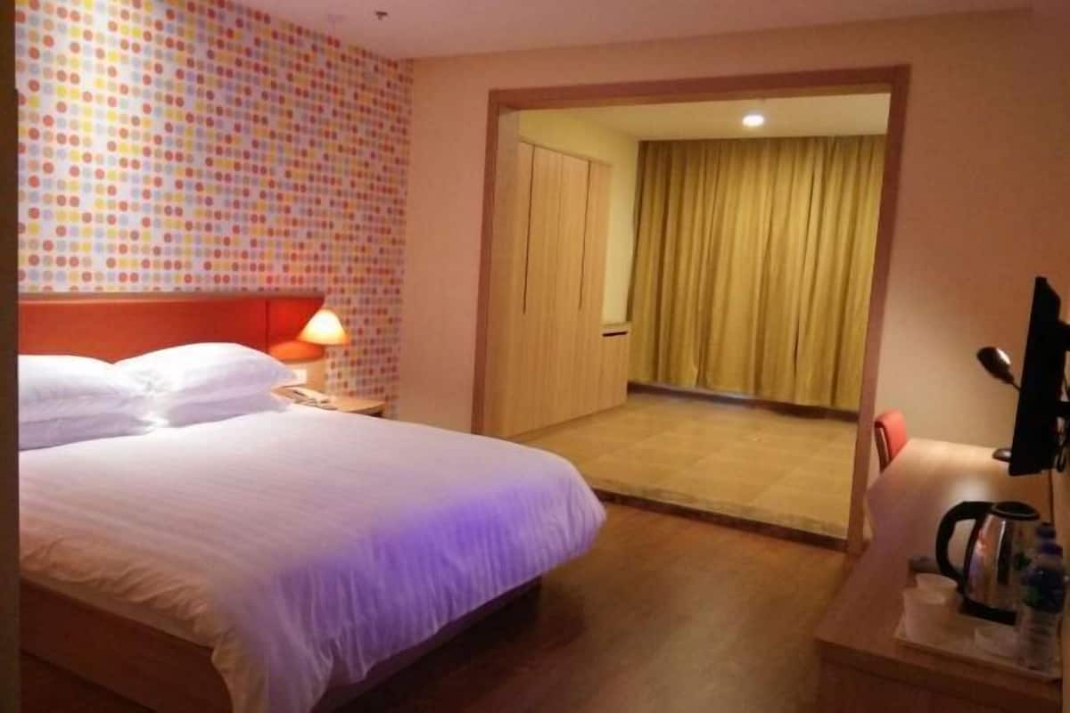 A hotel room with a bed, a nightstand with a lamp, a desk with a kettle and a bottle, and a door leading to a balcony.