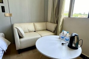 Hotellihuone, jossa on beigenvärinen sohva, pyöreä valkoinen pöytä, jossa on vedenkeitin ja vesipulloja, sekä ikkuna, jossa on verhot.