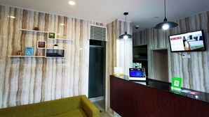 Un hall d’hôtel avec un mur décoré d’un motif imitant le marbre, un comptoir d’accueil, un téléviseur et un canapé jaune.