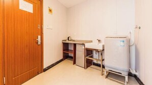 Une chambre d’hôtel de taille réduite, dotée d’une porte en bois, d’une petite kitchenette, d’un évier blanc et d’un réfrigérateur compact.