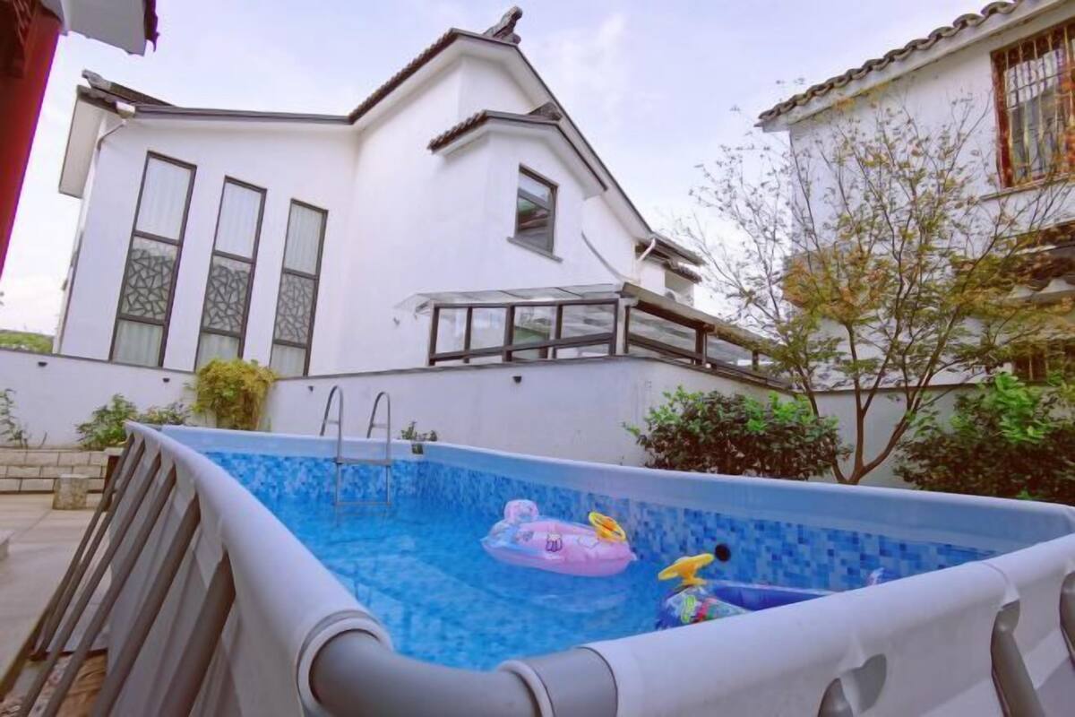 An outdoor swimming pool with inflatable toys, a ladder, and a white house with large windows and a tiled roof in the background.