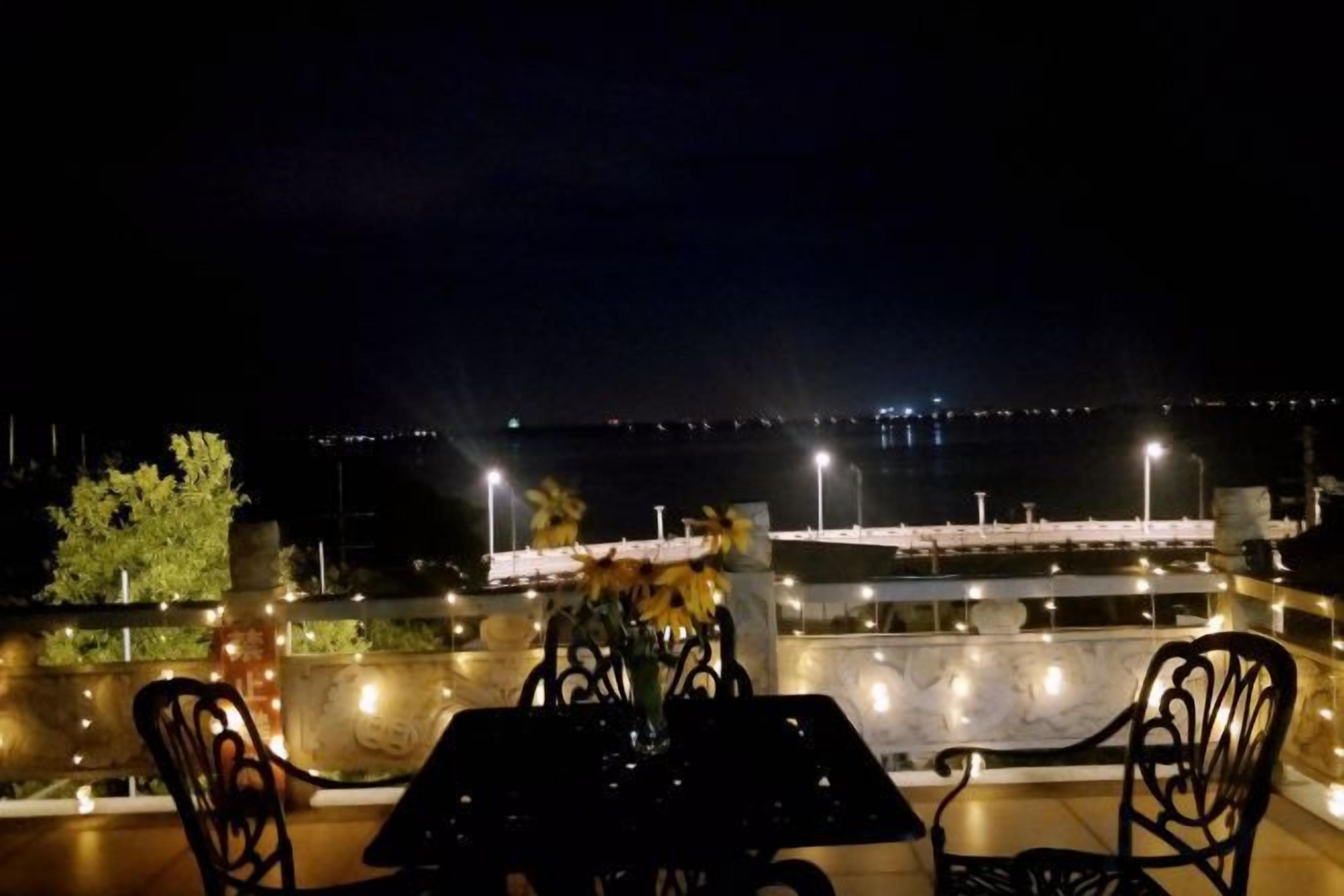 An outdoor dining area with a table and chairs, overlooking a waterfront with lights in the distance.