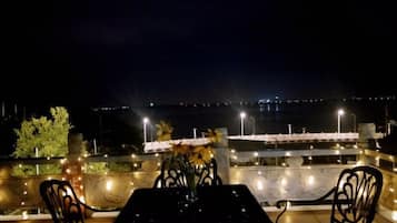 An outdoor dining area with a table and chairs, overlooking a waterfront with lights in the distance.