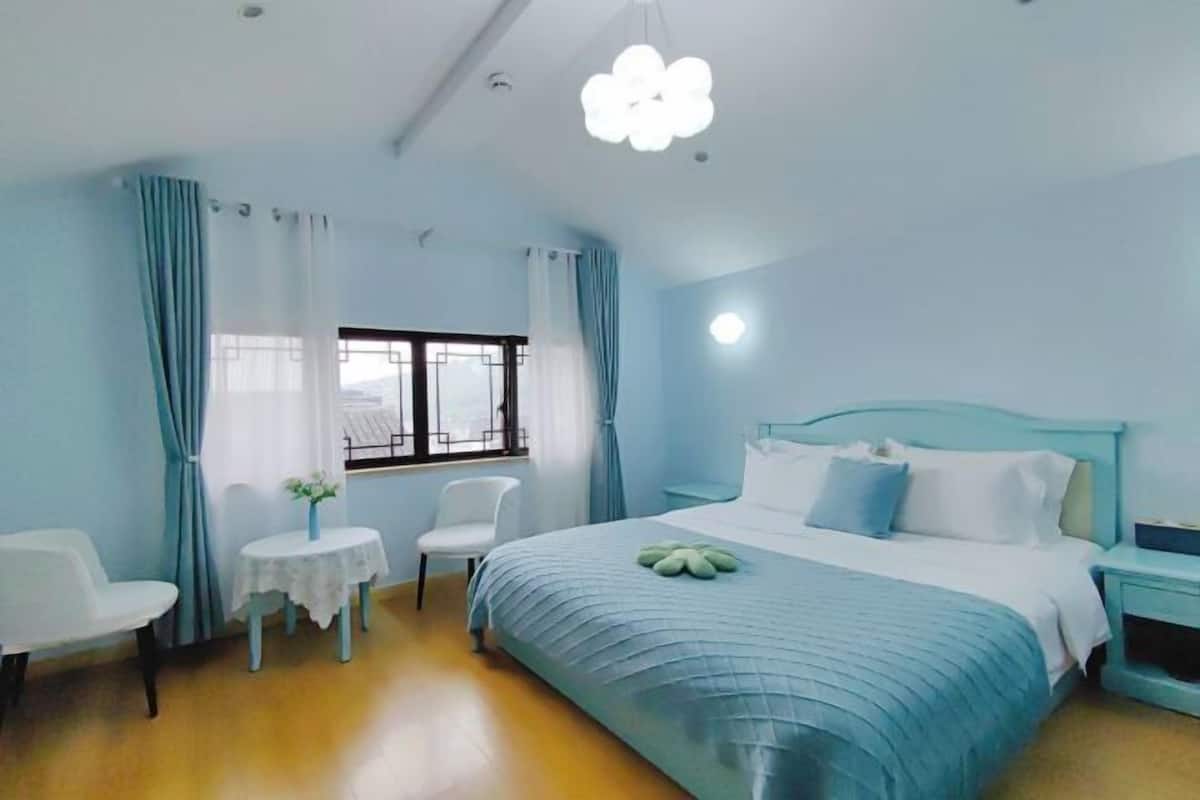 A bedroom with a bed, a bedside table, a chair, a small table with a vase, and a window with curtains.