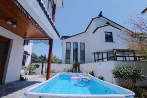 Een zwembad met drijvende speelgoed, een wit gebouw met schuine kap en een houten pergola.
