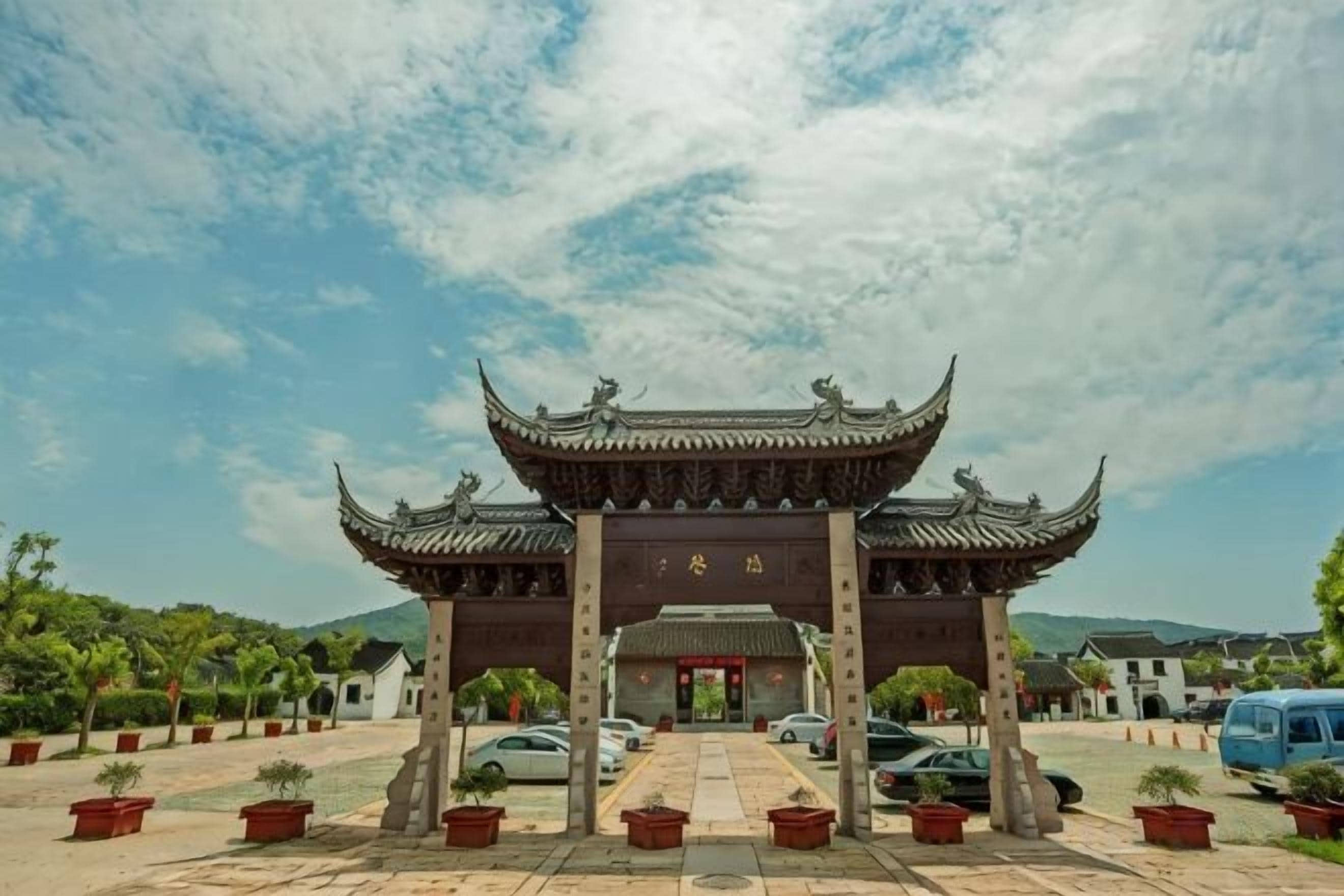 A traditional Chinese gate with a tiled roof and a red door, surrounded by potted plants and parked cars.