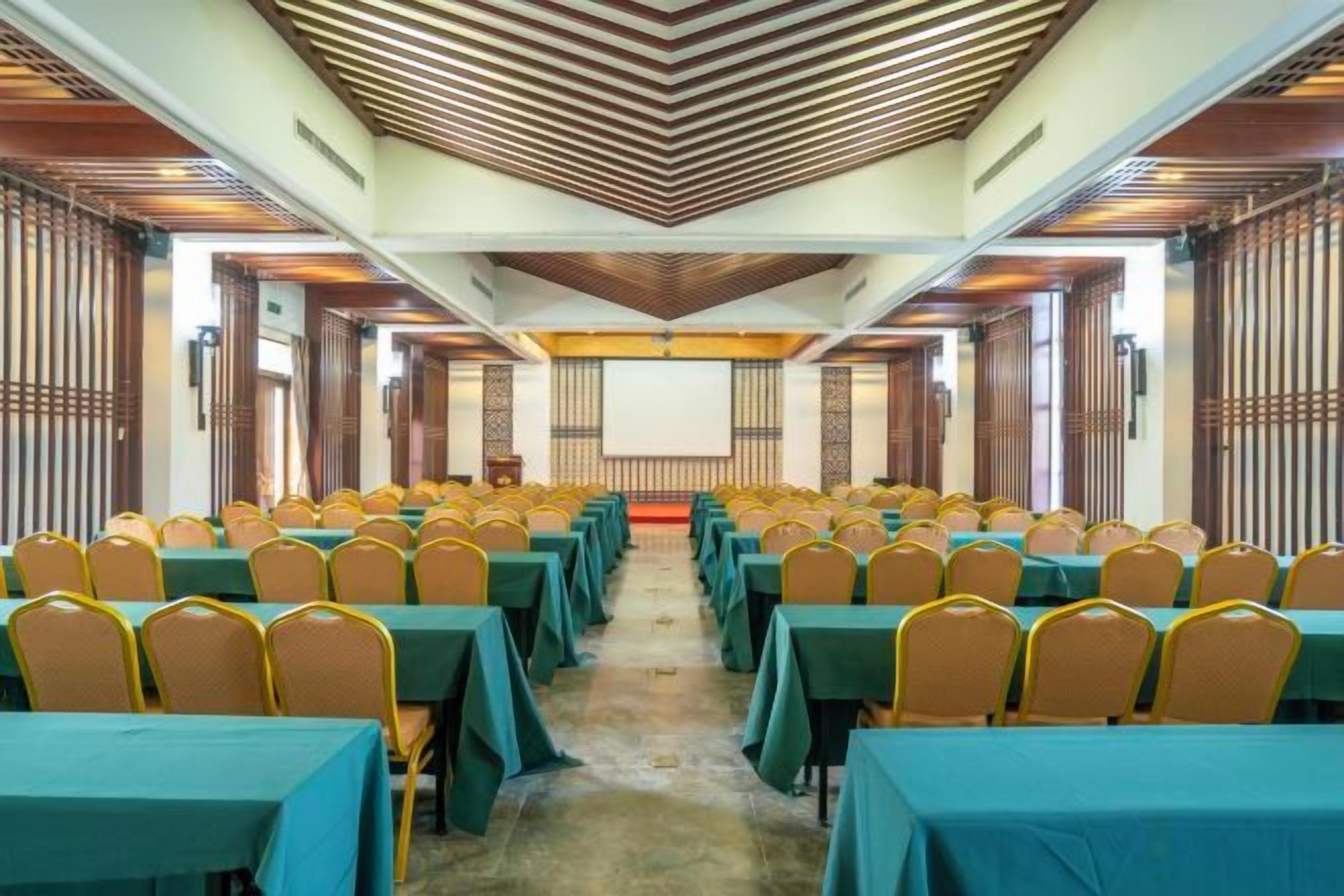 Un salón de conferencias con filas de mesas y sillas, techo de madera y una pantalla grande al frente.