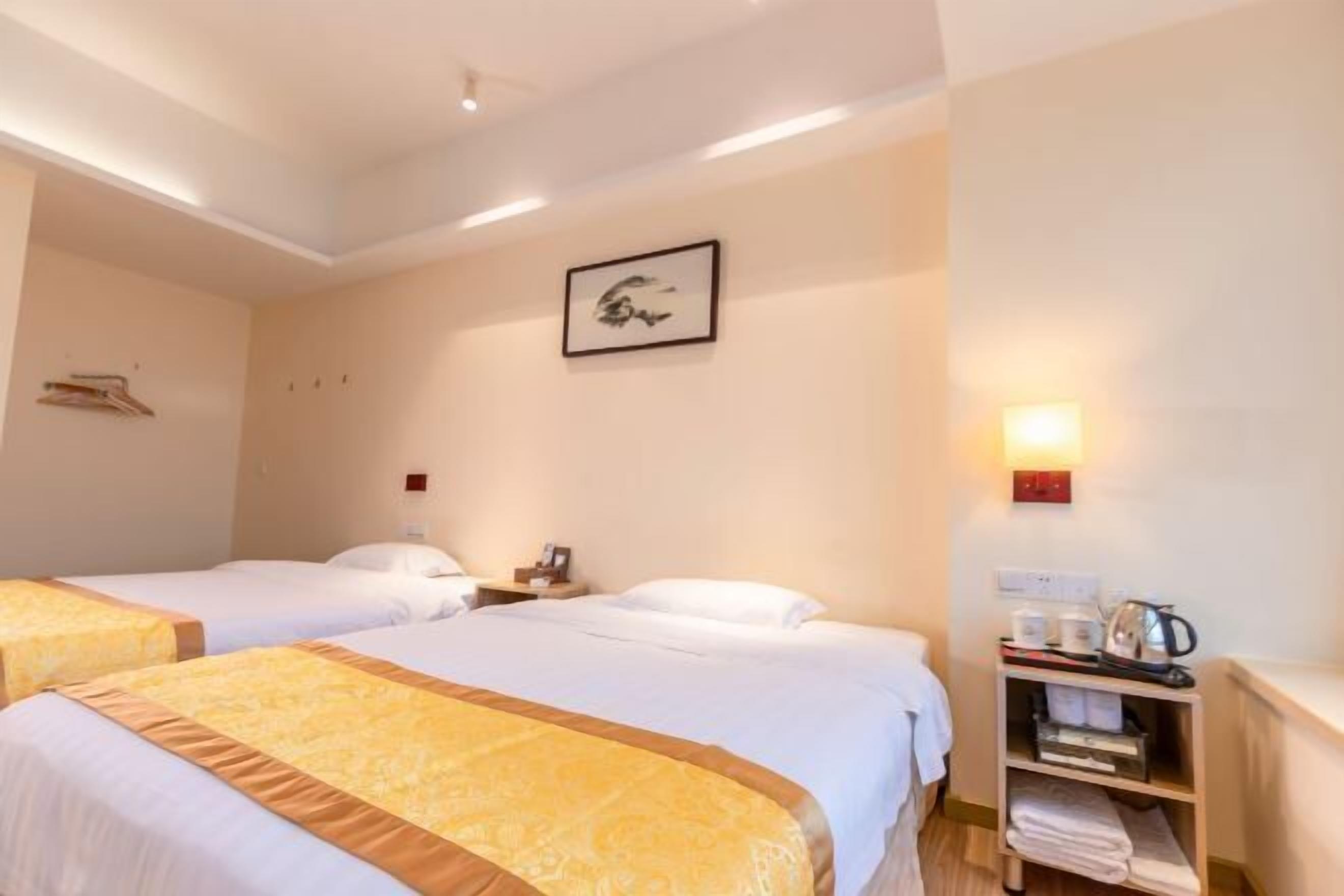 A hotel room with two beds, a nightstand, a wall-mounted light, and a shelf with towels and a kettle.