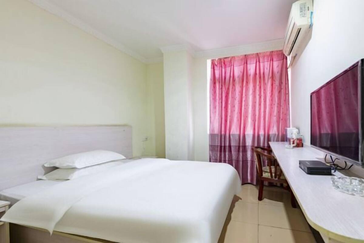 A hotel room with a bed, a desk, a chair, an air conditioning unit, and a television.