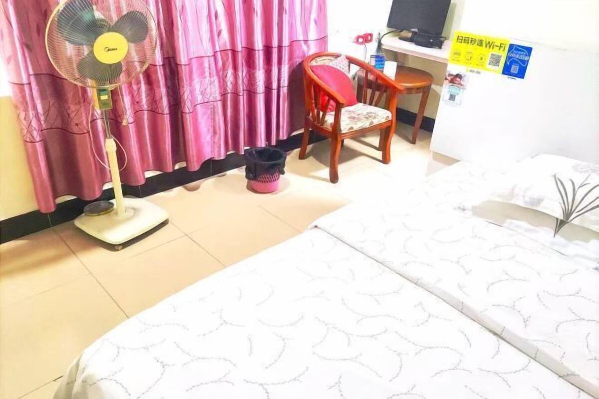 A hotel room with a bed, a standing fan, a chair, a desk, and a curtain.