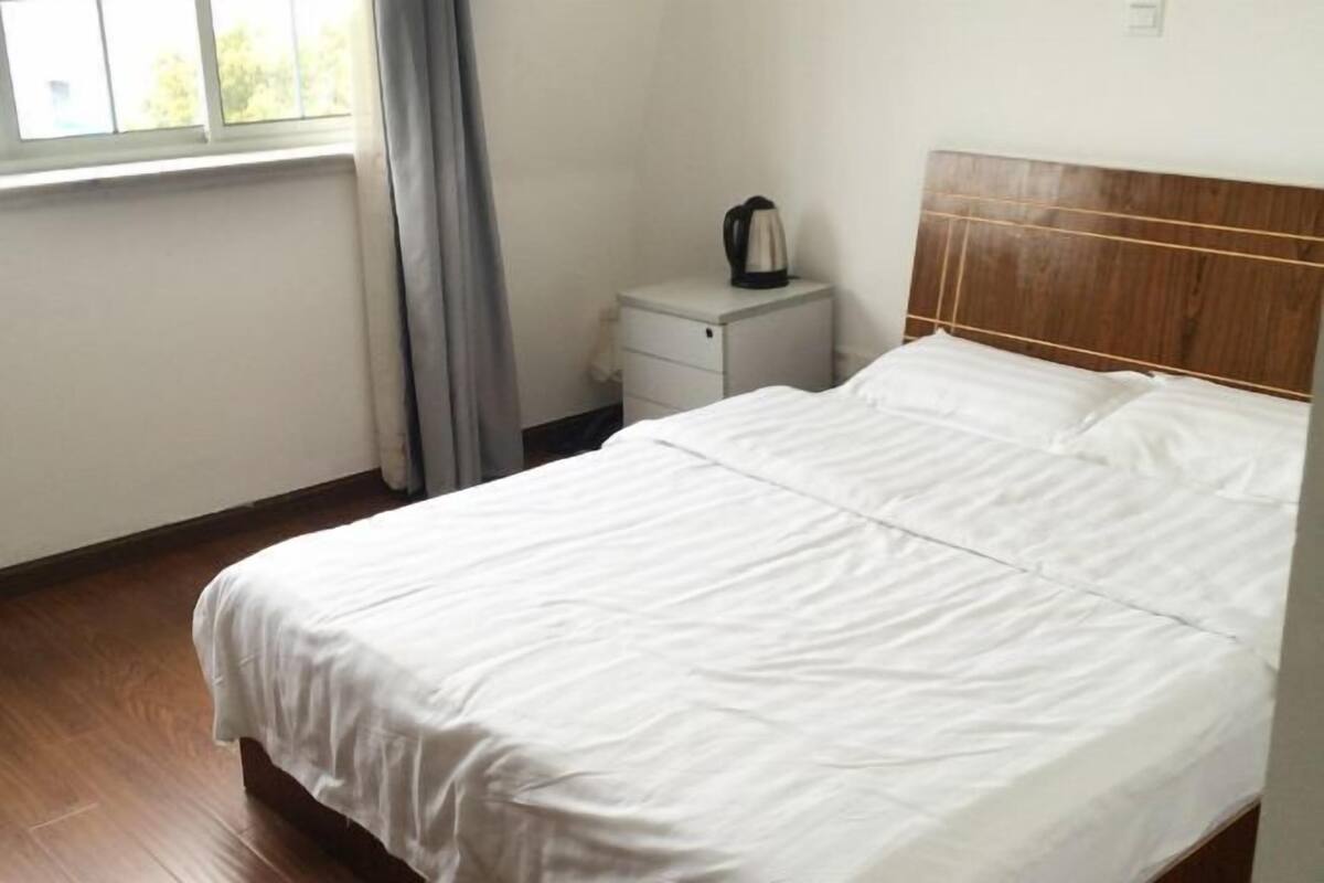 A hotel room with a wooden bed, a small bedside table with a kettle, and a window with curtains.