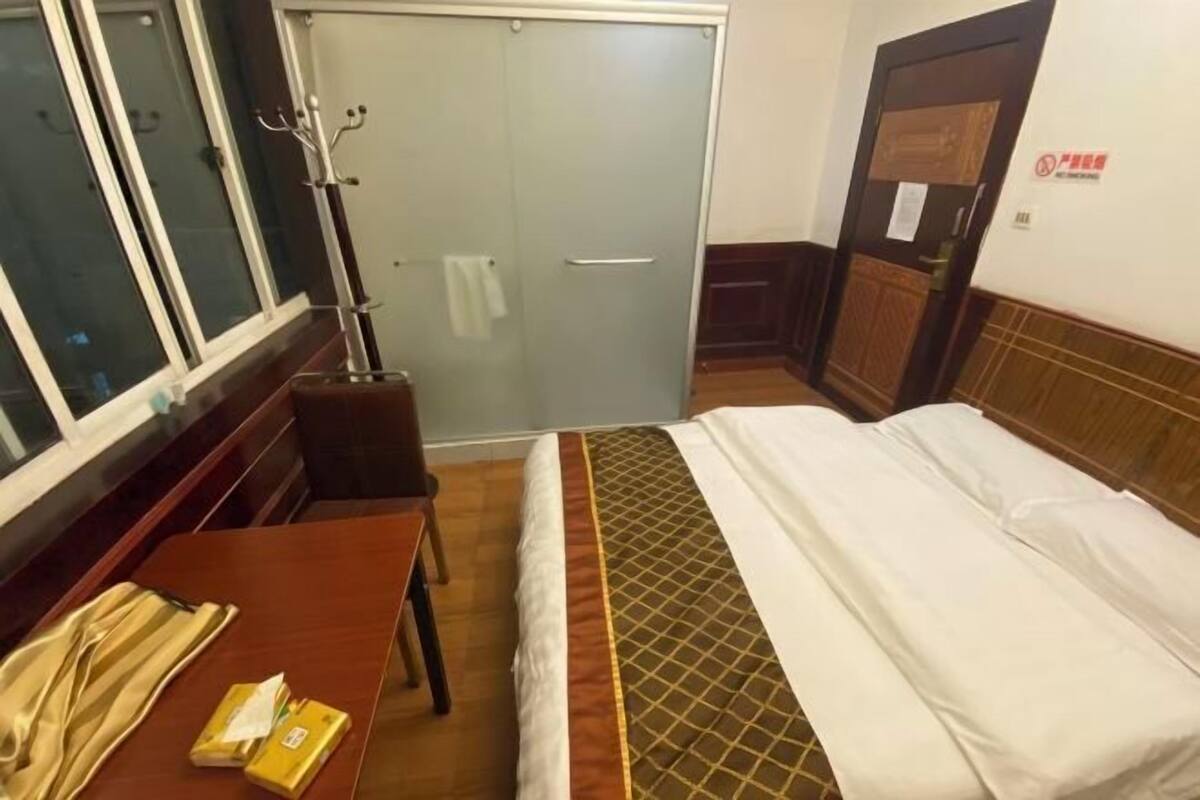 A hotel room with a bed, a wooden table with a telephone and a book, a window, a door, and a coat rack.