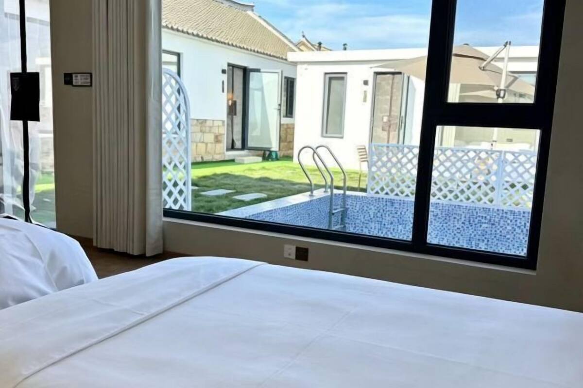 A hotel room with a large window, a bed, and a view of the outdoor swimming pool area.
